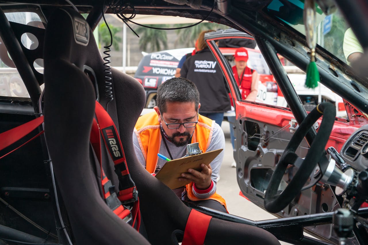 A man inspects a race car interior at a motorsport event, noting findings on a clipboard.