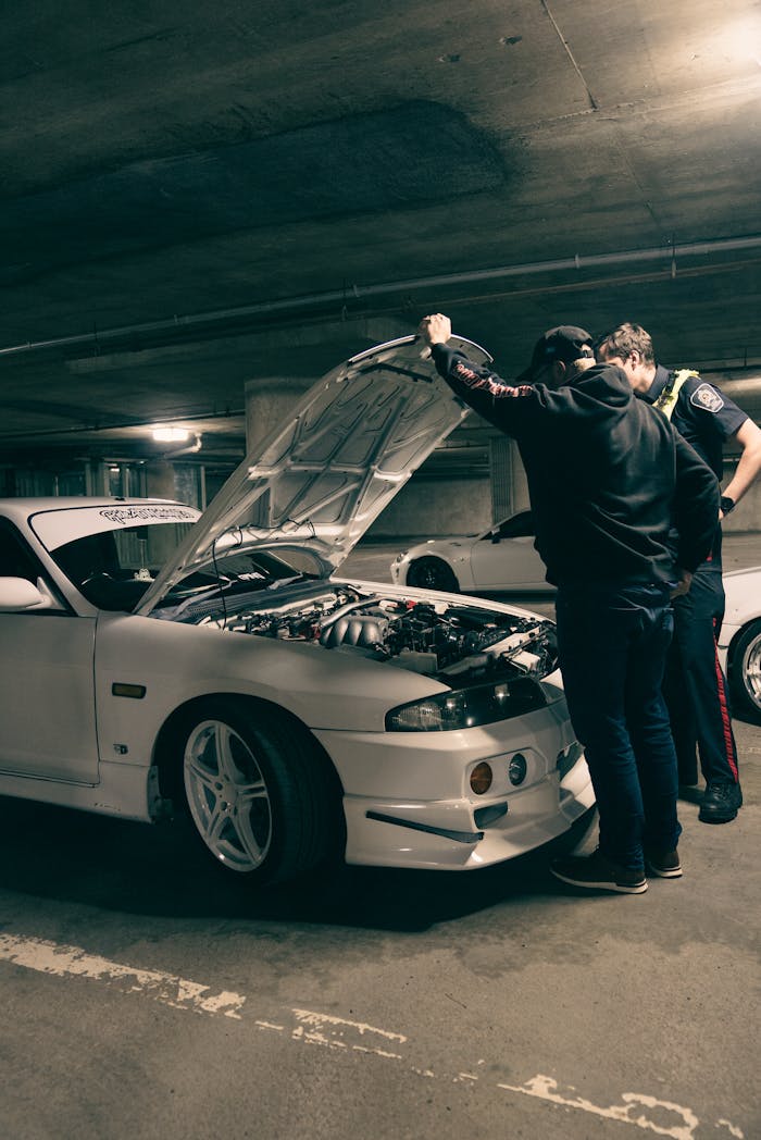 Two men examining a car engine in a dimly lit underground parking area.