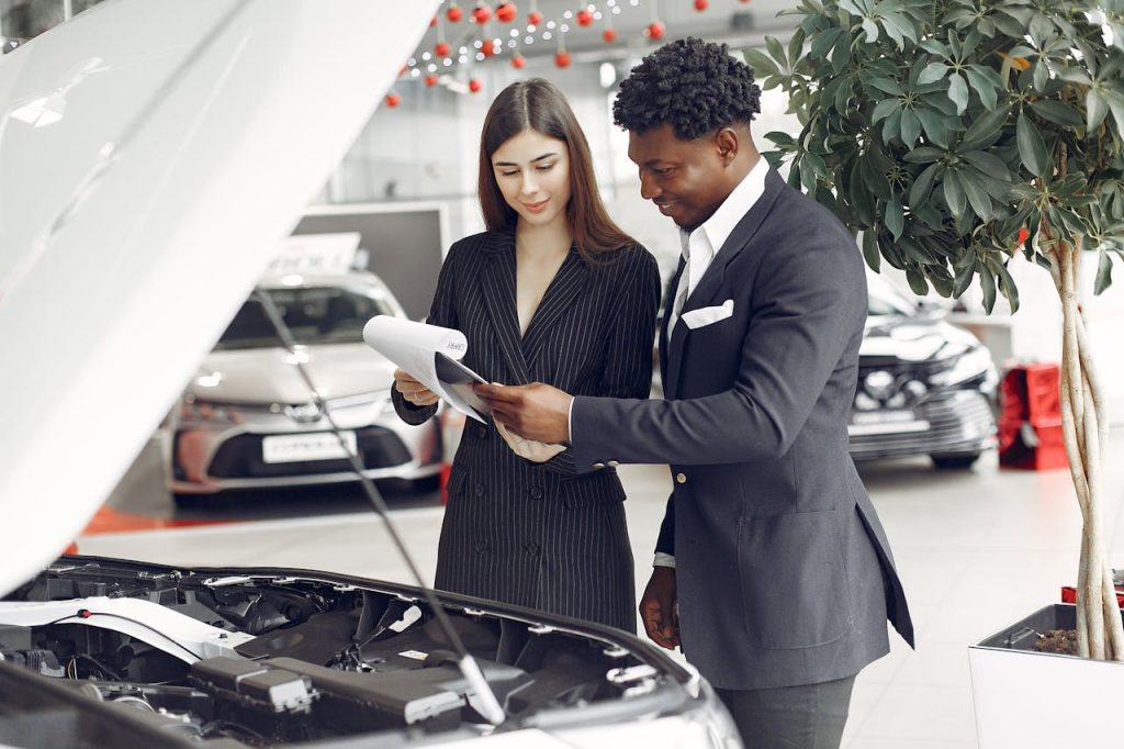 Confident smiling successful black businessman in classy formal suit standing near open motor hood in car showroom and discussing car characteristics with gorgeous female dealer